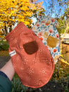 A hand holding two colorful, patterned dog vests against a backdrop of autumn foliage.