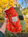 A person's hand is holding a red dog coat with a pattern of white bones, against a backdrop of autumn foliage.