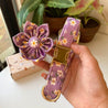 A hand holding a purple collar with a flower-shaped buckle and a potted plant in the background.