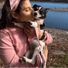 A woman in a pink sweater is affectionately kissing a small dog with a pink and white checkered bandana around its neck.