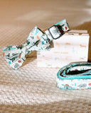 A white bow tie with a pattern of elephants and other animals, along with a matching white collar and leash, are displayed on a beige carpeted floor.