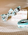 A white bow tie with a pattern of elephants and other animals, along with a matching white collar and leash, are displayed on a beige carpeted floor.